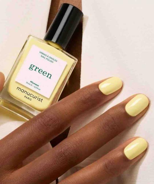 Manucurist Mimosa nail polish bottle held in a hand on tanned skin, with an elegant beige outfit.