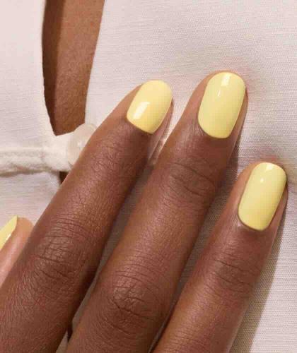 Gros plan sur une manucure jaune pastel Mimosa Manucurist sur une peau hâlée, avec un décolleté et une tenue beige élégante.