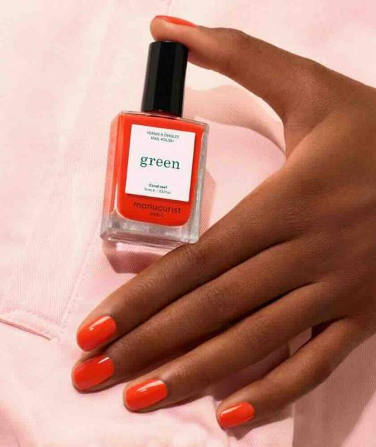 Coral Reef Manucurist manicure on tanned skin with the coral nail polish bottle on a pale pink background.