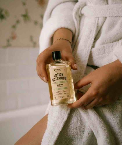 Lotion Botanique Körperöl von Féret Parfumeur, 100% natürlich, in den Händen einer Frau im weißen Bademantel in einem Badezimmer